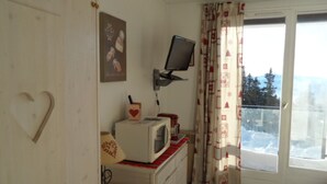 A small room with a white wall, a heart-shaped cutout, a microwave, a small table with a red and white checkered cloth, a television mounted on the wall, and a window with a view of snow-covered trees. - Studio cabin for 4 near slopes, Southwest exposure in Chamrousse (Chamrousse)