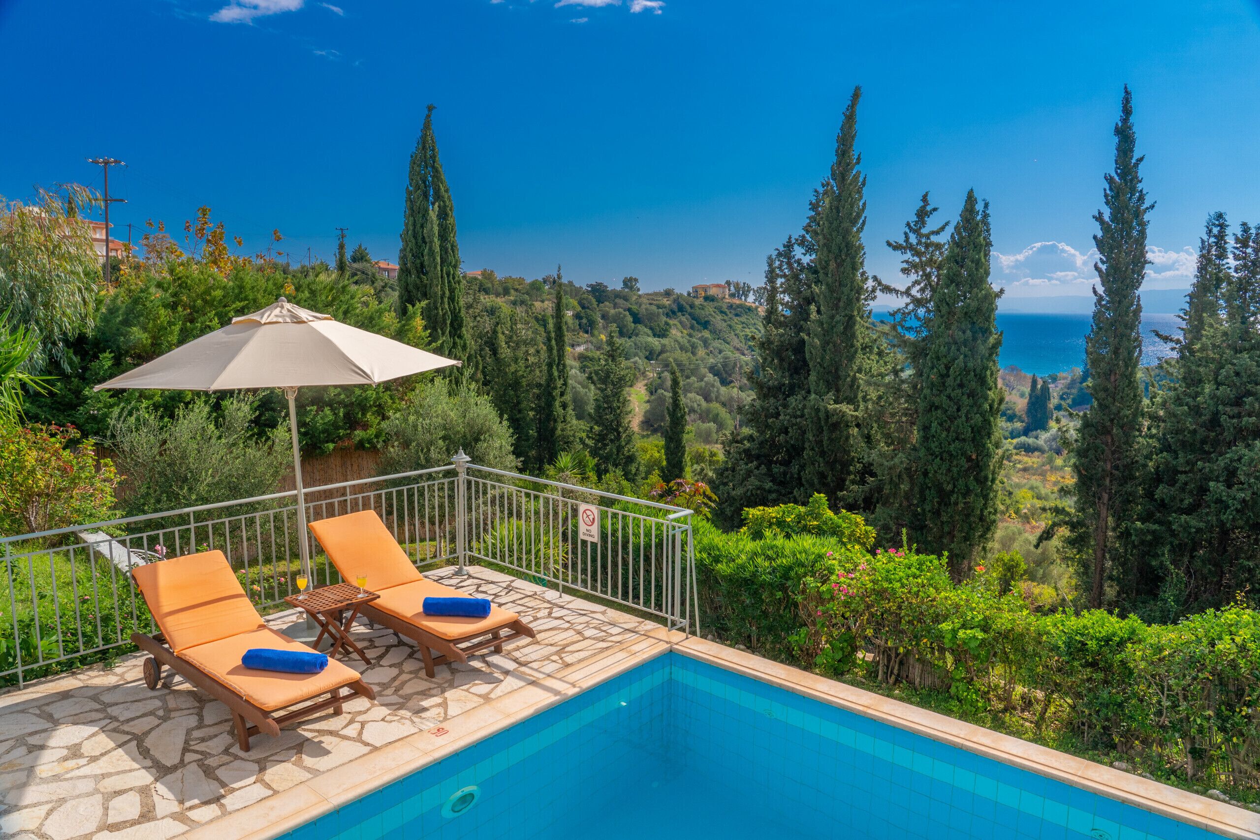 A poolside area with two lounge chairs and an umbrella, surrounded by greenery and overlooking a scenic view.