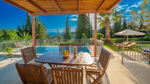 Un patio techado al aire libre con piscina, sillas de madera y una mesa con fruteros y copas de vino.