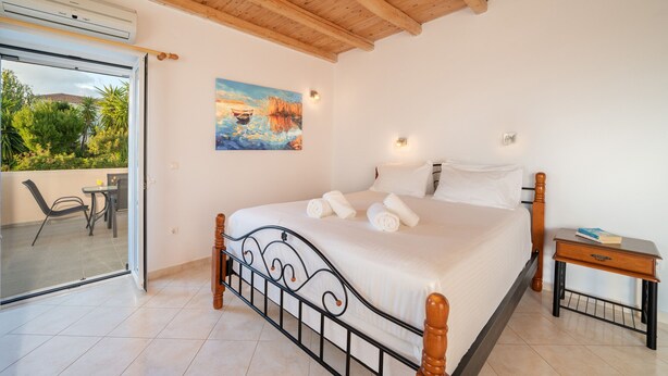 A bedroom with a bed, a painting, a nightstand, and a view of the outdoors.