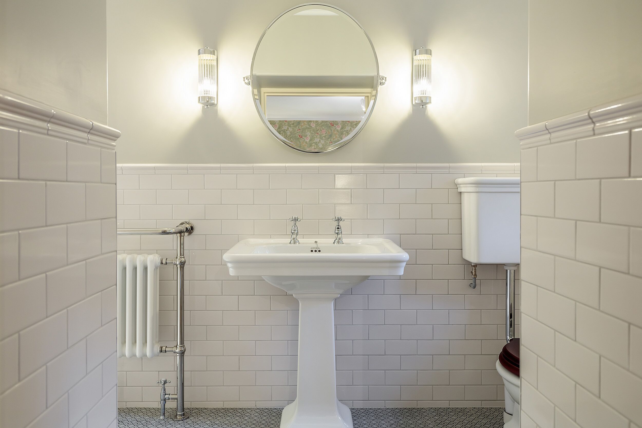 Un baño con lavamanos pedestal, un espejo redondo y dos luces fijadas a la pared.