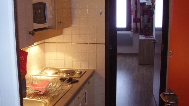 Kompakti keittiö, jossa on mikroaaltouuni, liesi ja astianpesukone. Punainen pyyhe on ripustettu uunin oveen. Keittiöstä on kulku eteiseen, jossa on ikkuna ja ovi toiseen huoneeseen.