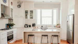 Una cocina moderna con armarios blancos, un refrigerador de acero inoxidable, una estufa a gas y un amplio ventanal que proporciona luz natural.