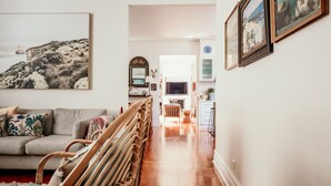 Una sala de estar acogedora con un sofá, un sillón de mimbre y un cuadro grande de un paisaje costero. Un pasillo conduce a una zona de cocina con un televisor y un reloj en la pared.