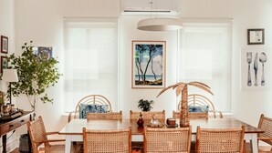Un comedor con una mesa de madera y sillas de ratán, un ventanal grande y un cuadro con árboles.