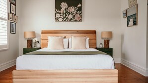 Un dormitorio con cabecera de madera, una cama grande con ropa blanca, dos mesitas de noche con lámparas, y una obra de arte floral sobre la cama.