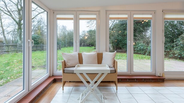A room with a large window, a wicker sofa with cushions, and a small table. - Vacation home Heidehaus for up to 6 people (Enge-Sande)