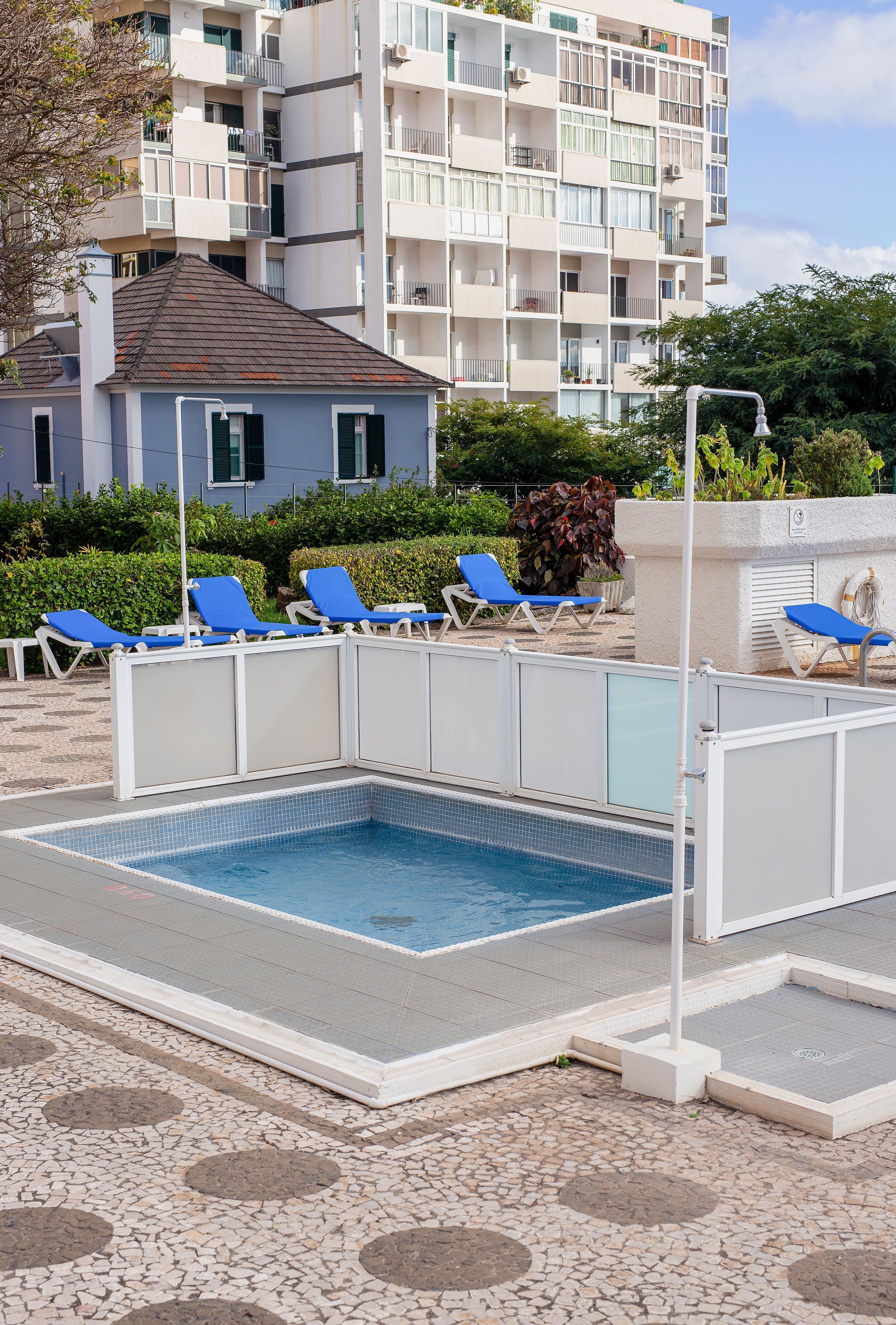 A swimming pool with blue lounge chairs, a white pool fence, and a building with balconies in the background.