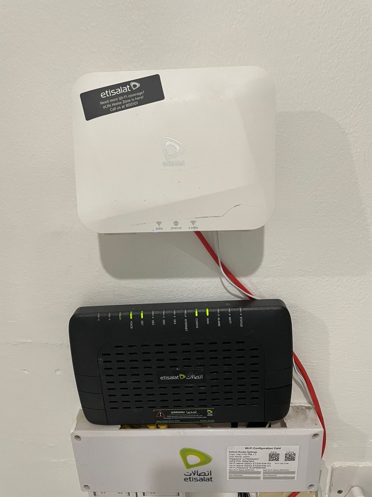 A white etisalat Wi-Fi router mounted on a white wall, connected to a black etisalat modem with a red and white cable.