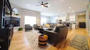 Ein modernes Wohnzimmer mit einer Couch, Sesseln, einem Couchtisch und einem Kamin. Im Hintergrund ist eine Küche mit Schränken und Geräten zu sehen.