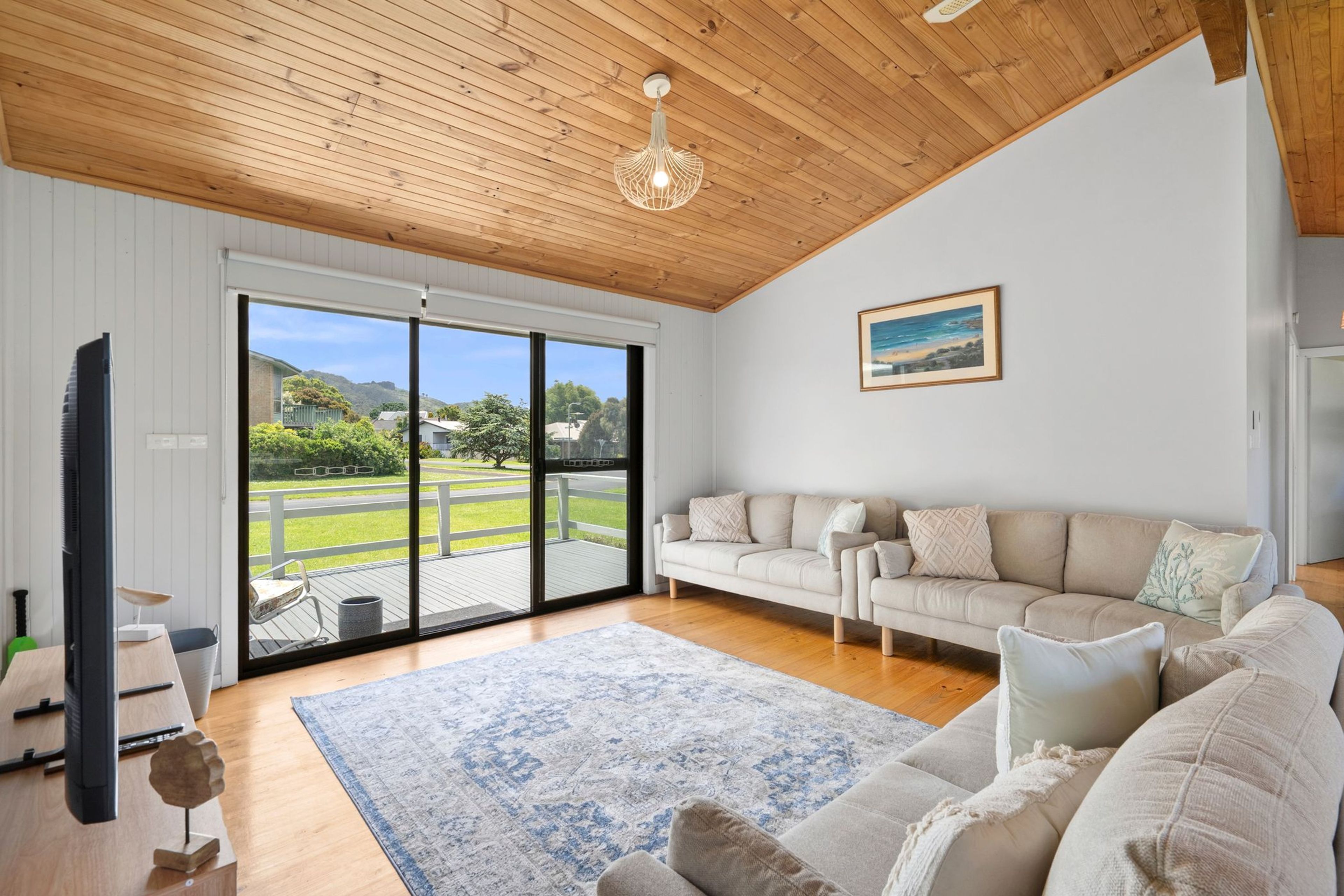 A spacious living room with a large sliding glass door leading to a deck, a flat-screen TV, a wooden ceiling, and a painting on the wall.