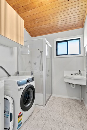 A laundry room with a front-loading washing machine, a glass-enclosed shower, and a window. - Shell Shack (Apollo Bay)