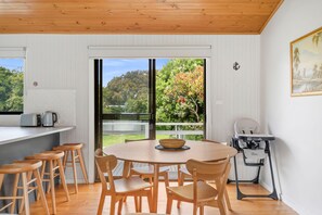 A dining area with a round table, wooden chairs, and a view of a garden through a glass door. - Shell Shack (Apollo Bay)