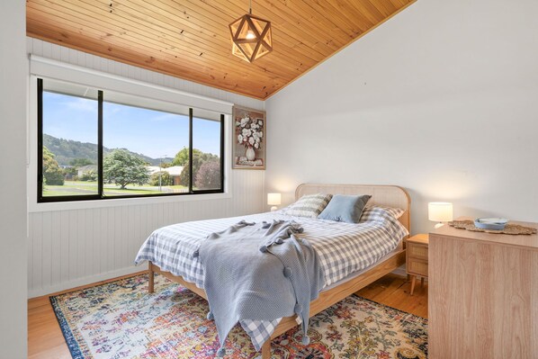 Une chambre avec une grande fenêtre, un lit recouvert d’une couvre-lit à carreaux, une tête de lit en bois, une table de chevet avec une lampe et un élément décoratif mural.