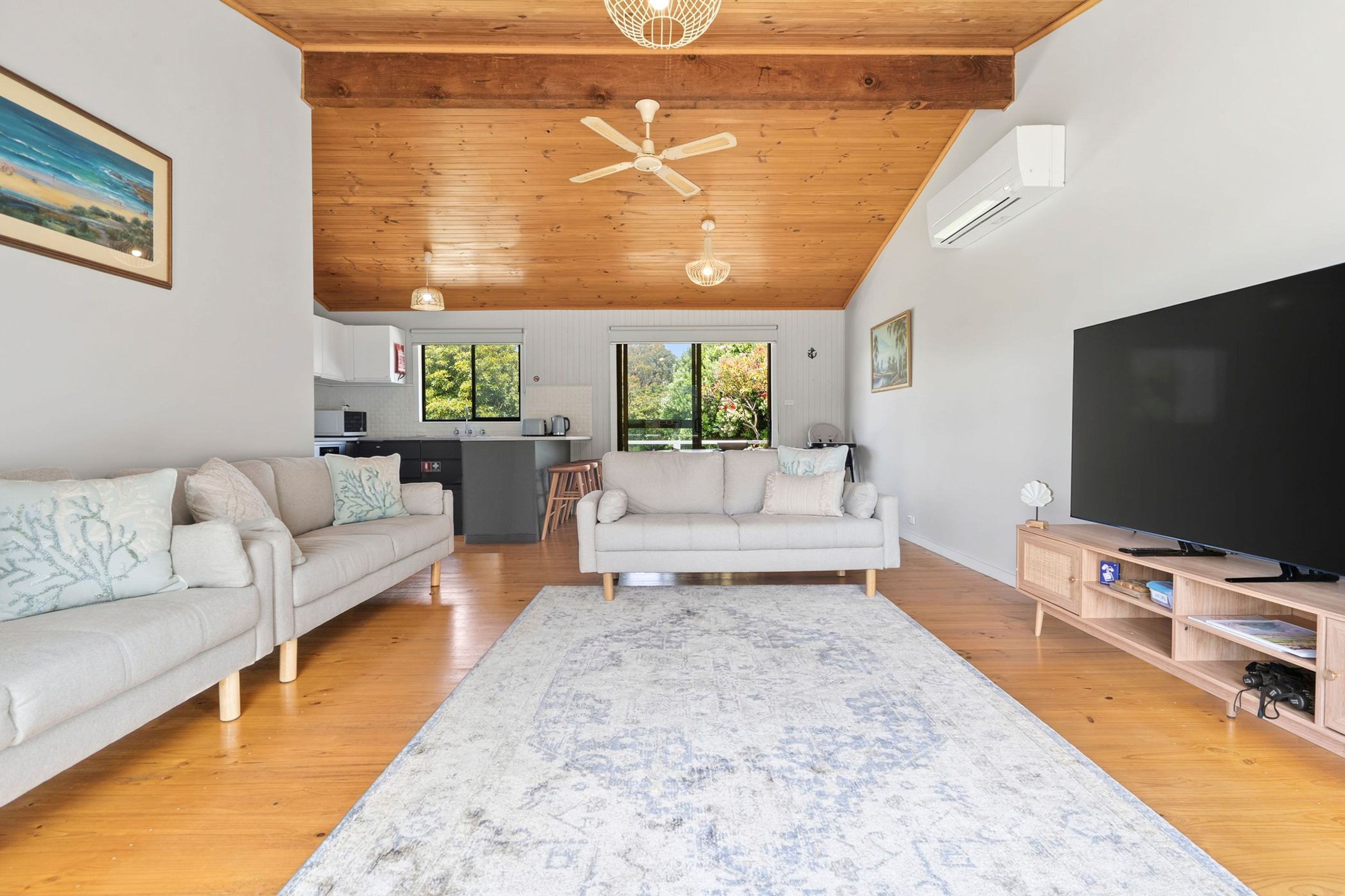 A spacious living area with a wooden ceiling, a large flat-screen TV, two sofas, a kitchen in the background, and a view of greenery outside.