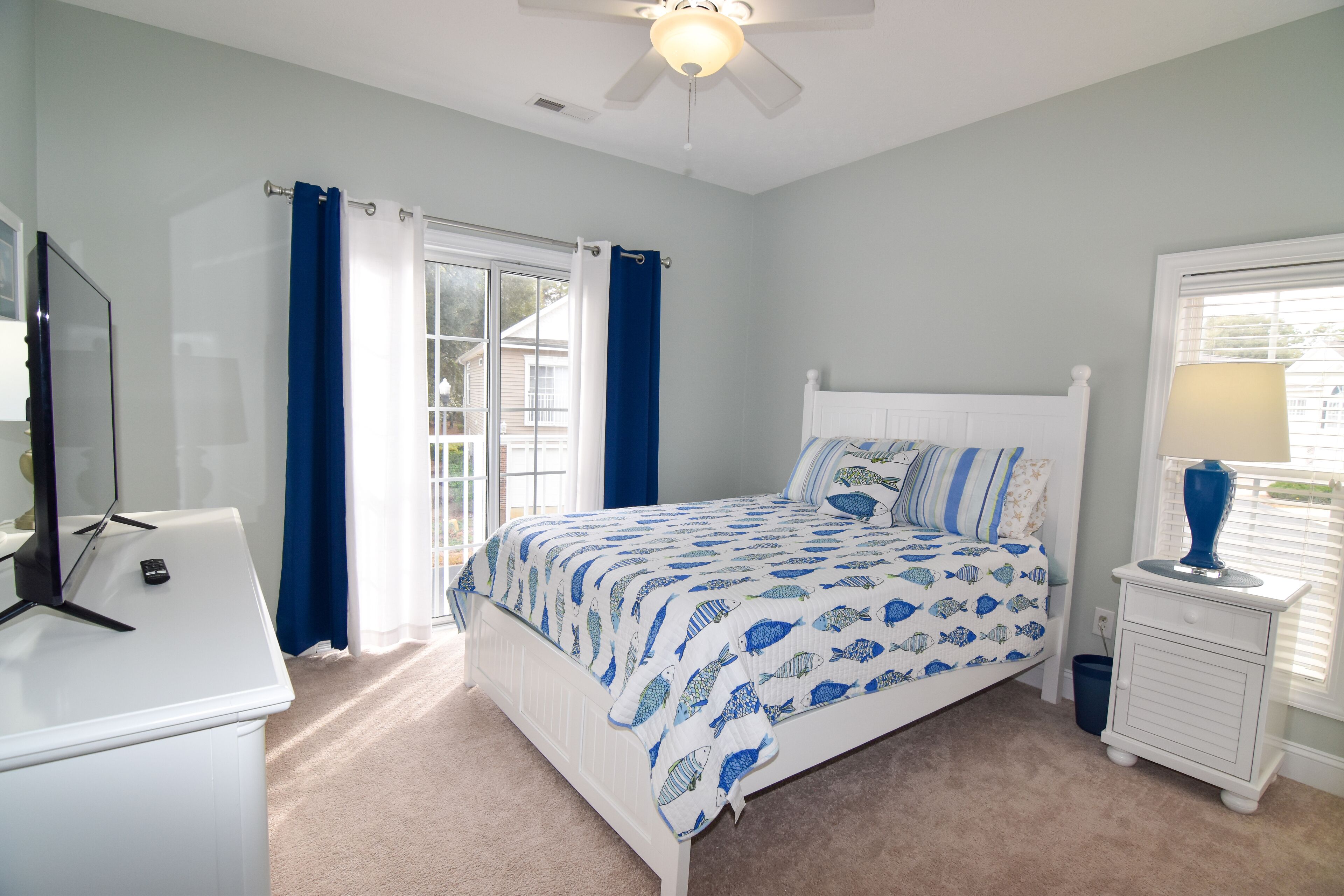 Une chambre à coucher comprenant un lit, une commode, une table de chevet, un ventilateur de plafond et une fenêtre avec des rideaux.