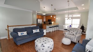 Ein geräumiges Wohnzimmer mit blauem Sofa, Hocker und einem gestreiften Sessel, im Hintergrund eine Küche und eine Treppe, die zu einer weiteren Ebene führt.