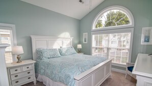 Ein Schlafzimmer mit einem großen Bett, weißen Nachttischen, einer Kommode und einem Fenster mit Jalousien.