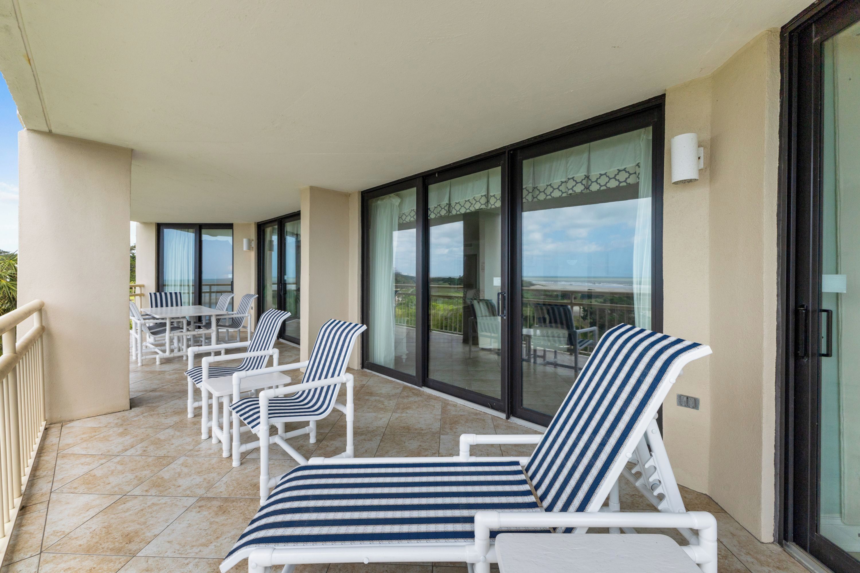 A balcony with white lounge chairs and a table, large glass doors leading to a terrace, and a clear view of the ocean.