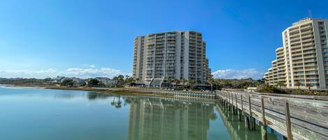 A waterfront view with two high-rise buildings, a calm body of water, and a wooden pier.