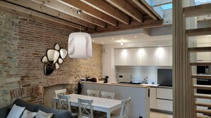 A modern kitchen with white cabinets and a central island, a dining area with a white table and chairs, and a living room with a grey sofa and decorative pillows.