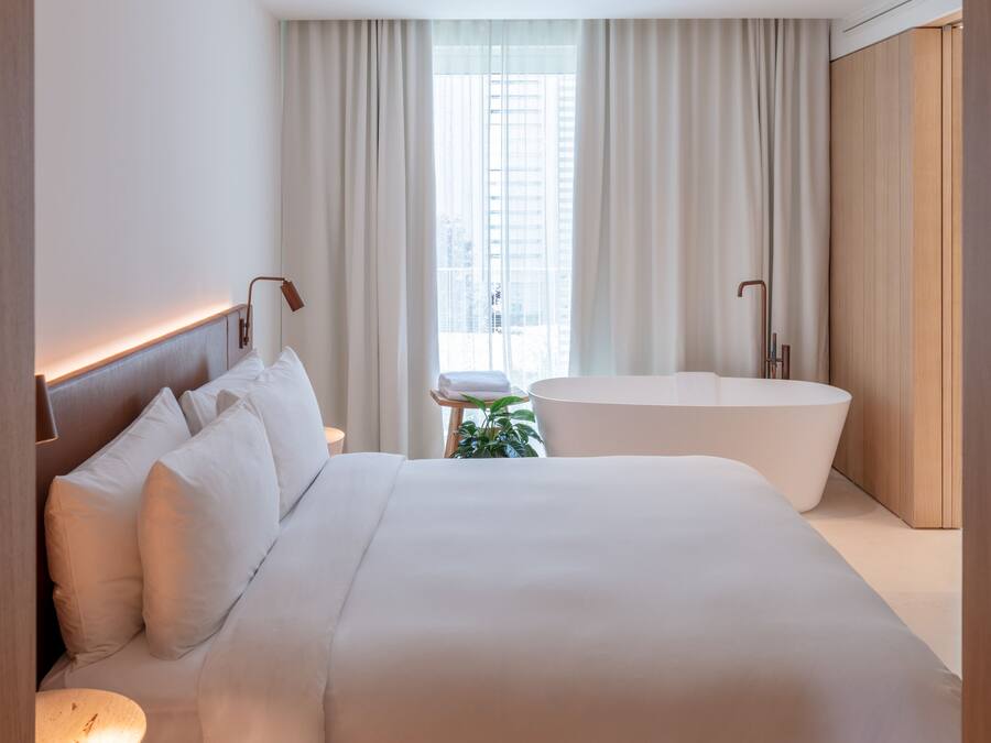 Une chambre d’hôtel moderne dotée d’un grand lit, d’une table de chevet, d’une baignoire intégrée et d’une fenêtre avec des rideaux.