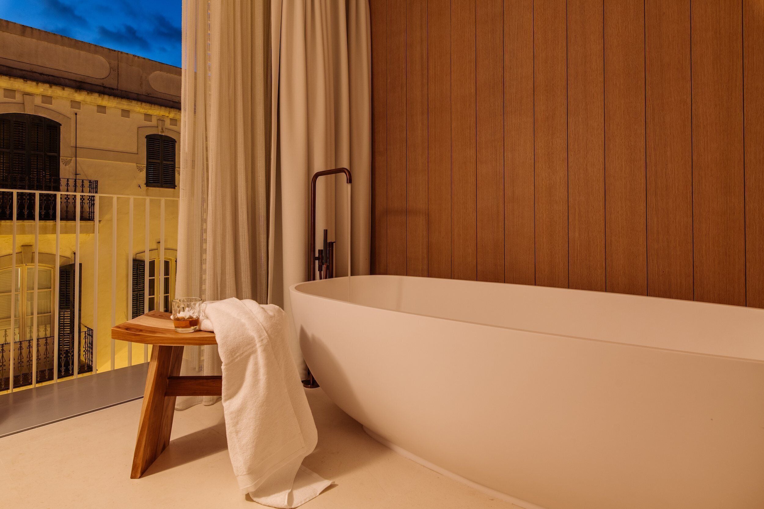 A modern bathroom with a freestanding bathtub, a wooden stool with a towel, and a view of a balcony with a railing.