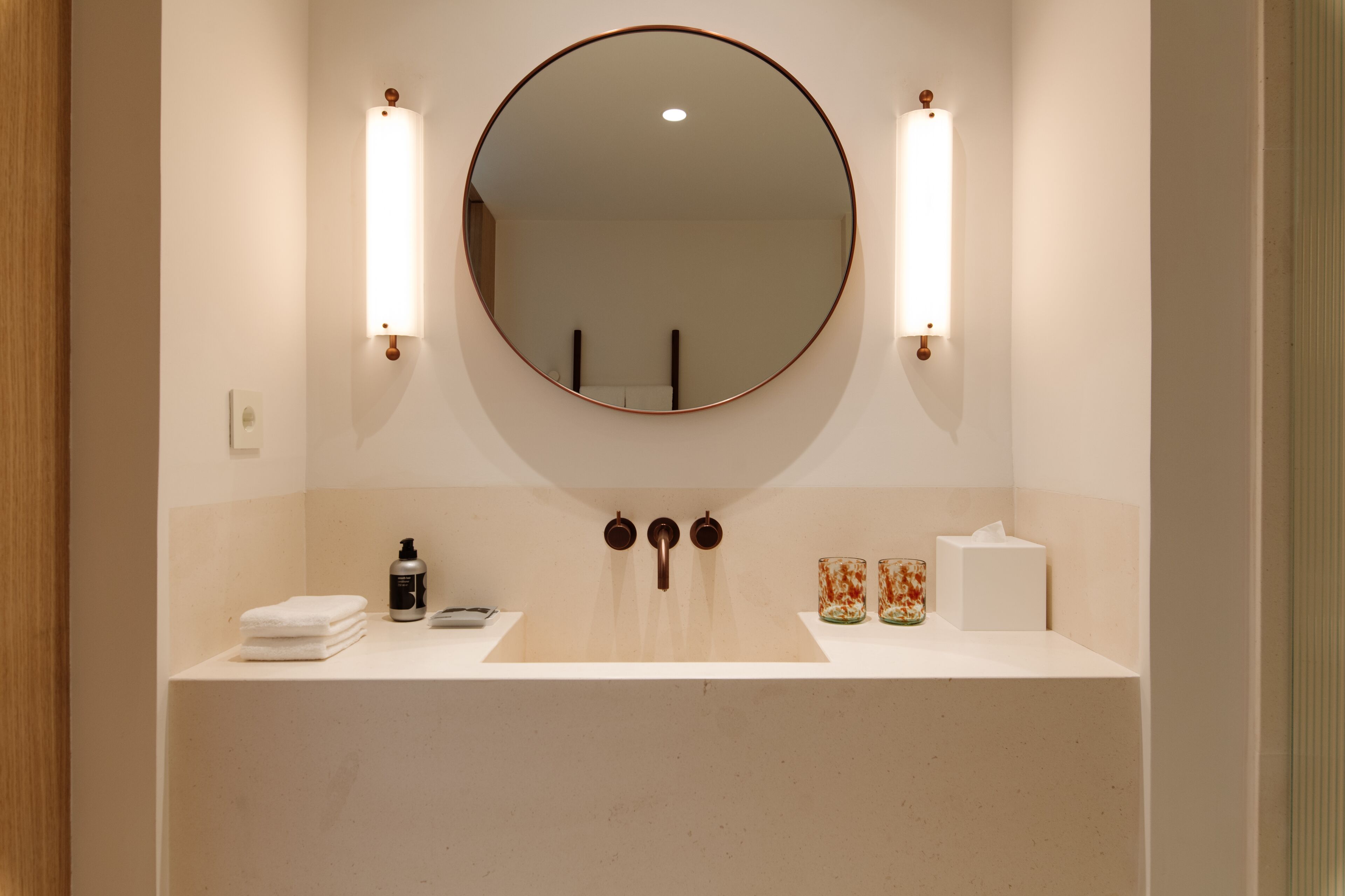 A bathroom with a round mirror, two wall-mounted lights, a sink, and a towel stack.
