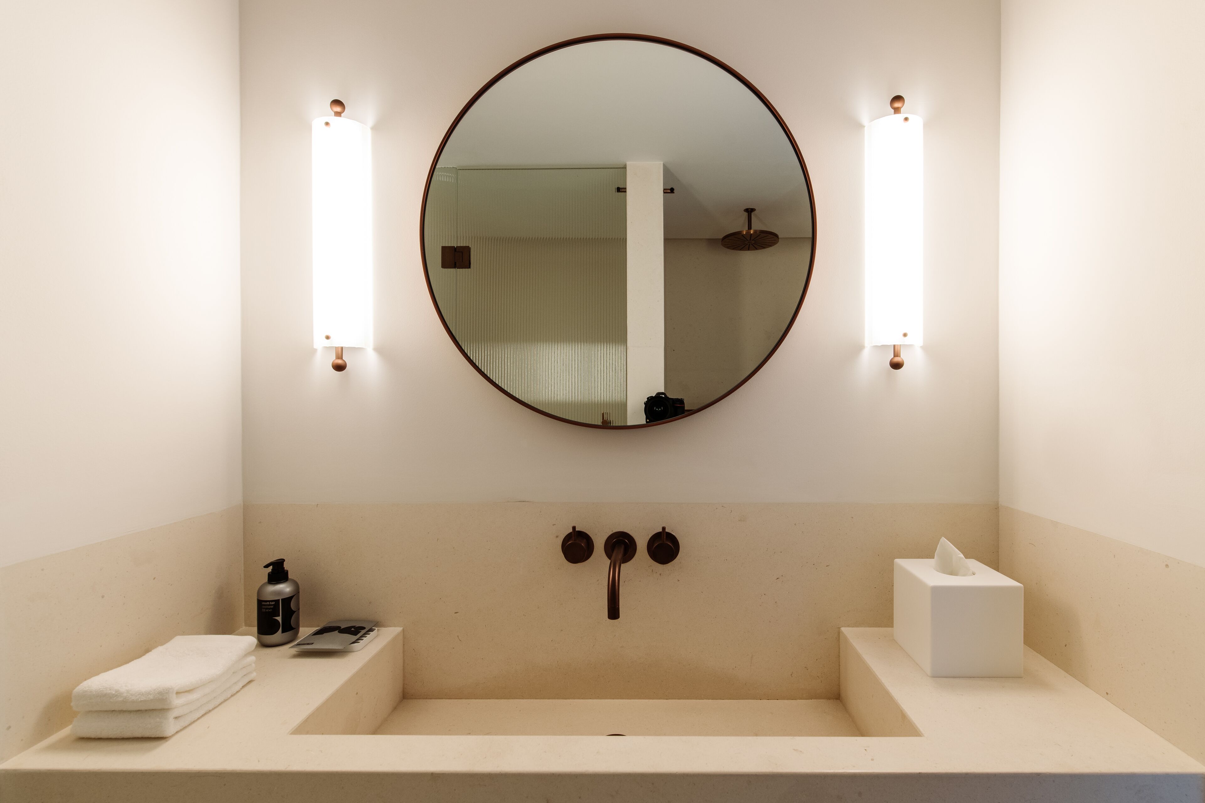 Un baño con un espejo redondo, dos luces empotradas, un lavabo, una toalla, un dispensador de jabón y una caja de pañuelos.