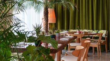 A restaurant with wooden tables and chairs, a palm tree, and green curtains.