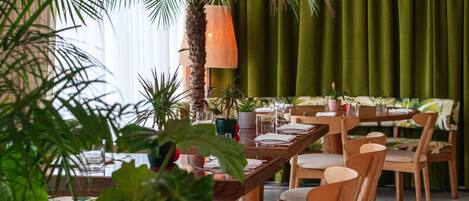 A restaurant with wooden tables and chairs, a palm tree, and green curtains.
