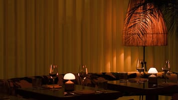Un restaurante tenuemente iluminado con mesas dispuestas para cenar, con un mantel estampado y una lámpara de hojas de palma.