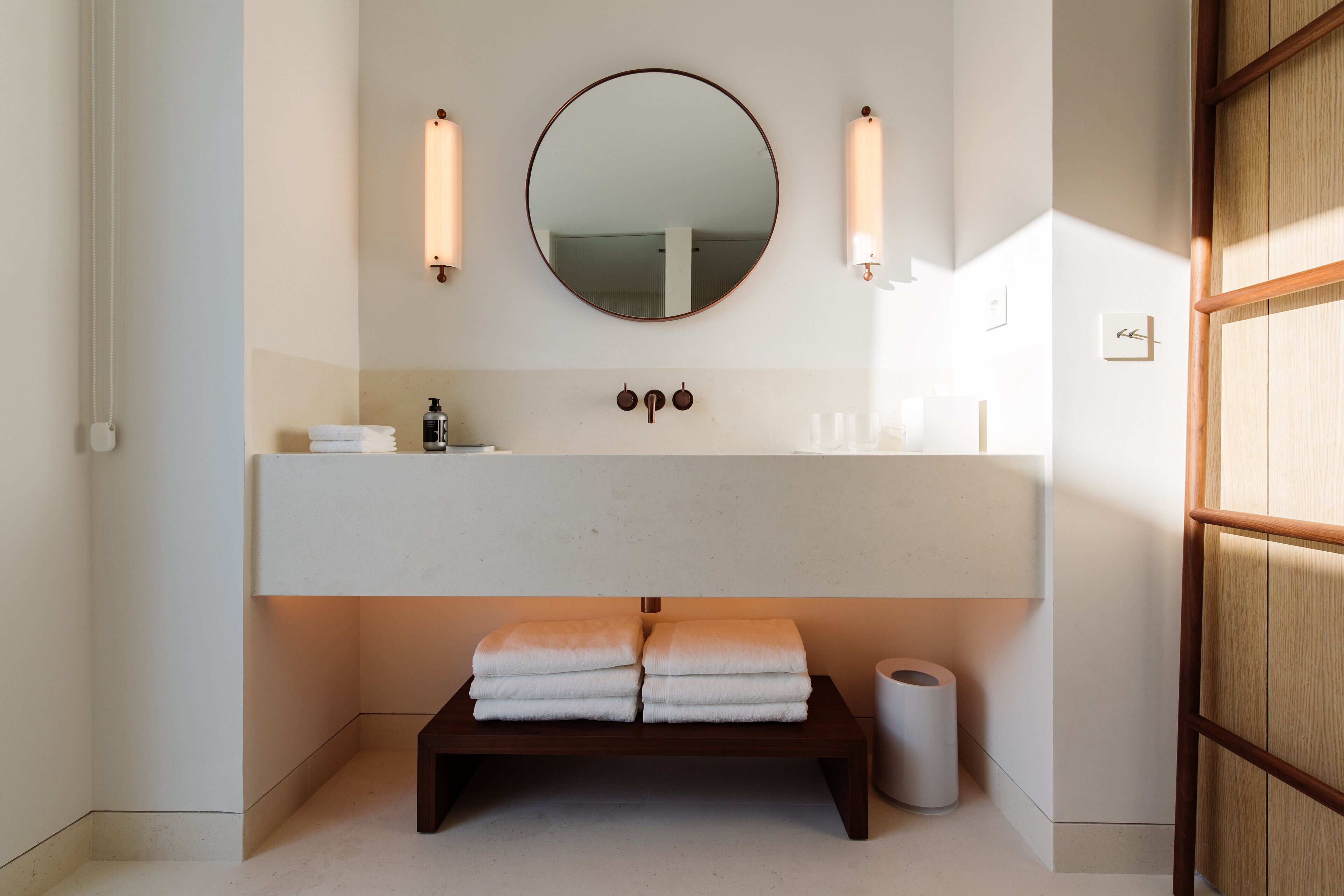 A bathroom with a large mirror, two wall-mounted lights, a wooden bench with towels, and a toilet.