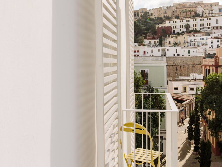 A balcony with a white railing and a yellow chair, overlooking a cityscape with buildings and a clear sky.