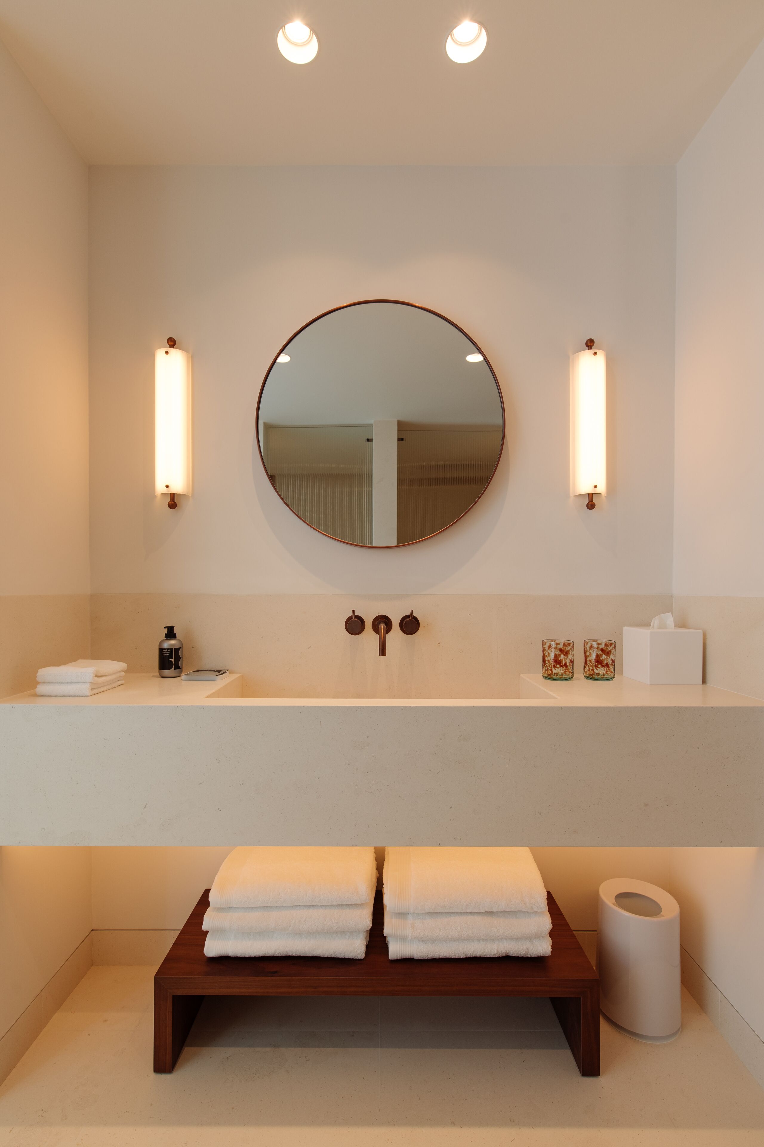 A modern bathroom with a round mirror, two wall-mounted lights, a wooden bench with towels, a sink, and a toilet paper holder.