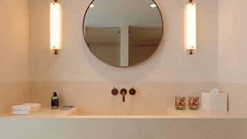 A modern bathroom with a round mirror, two wall-mounted lights, a wooden bench with towels, a sink, and a toilet paper holder.