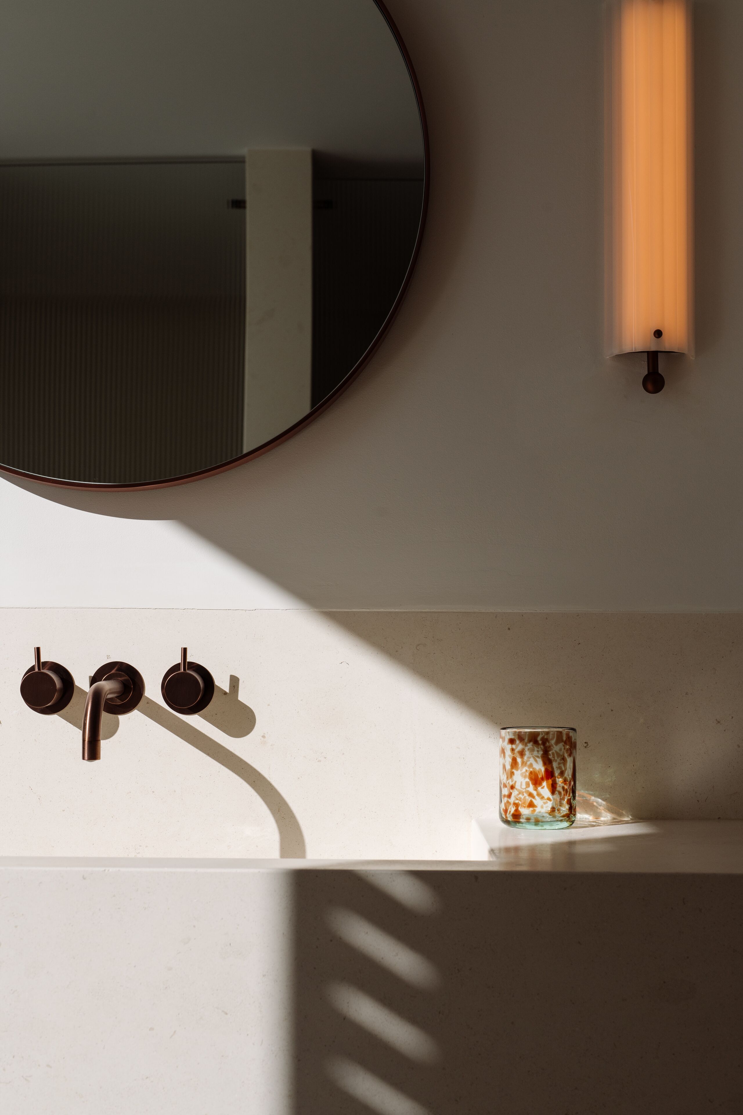 Un baño con un espejo redondo, una lámpara de pared y un lavamanos con tres grifos.