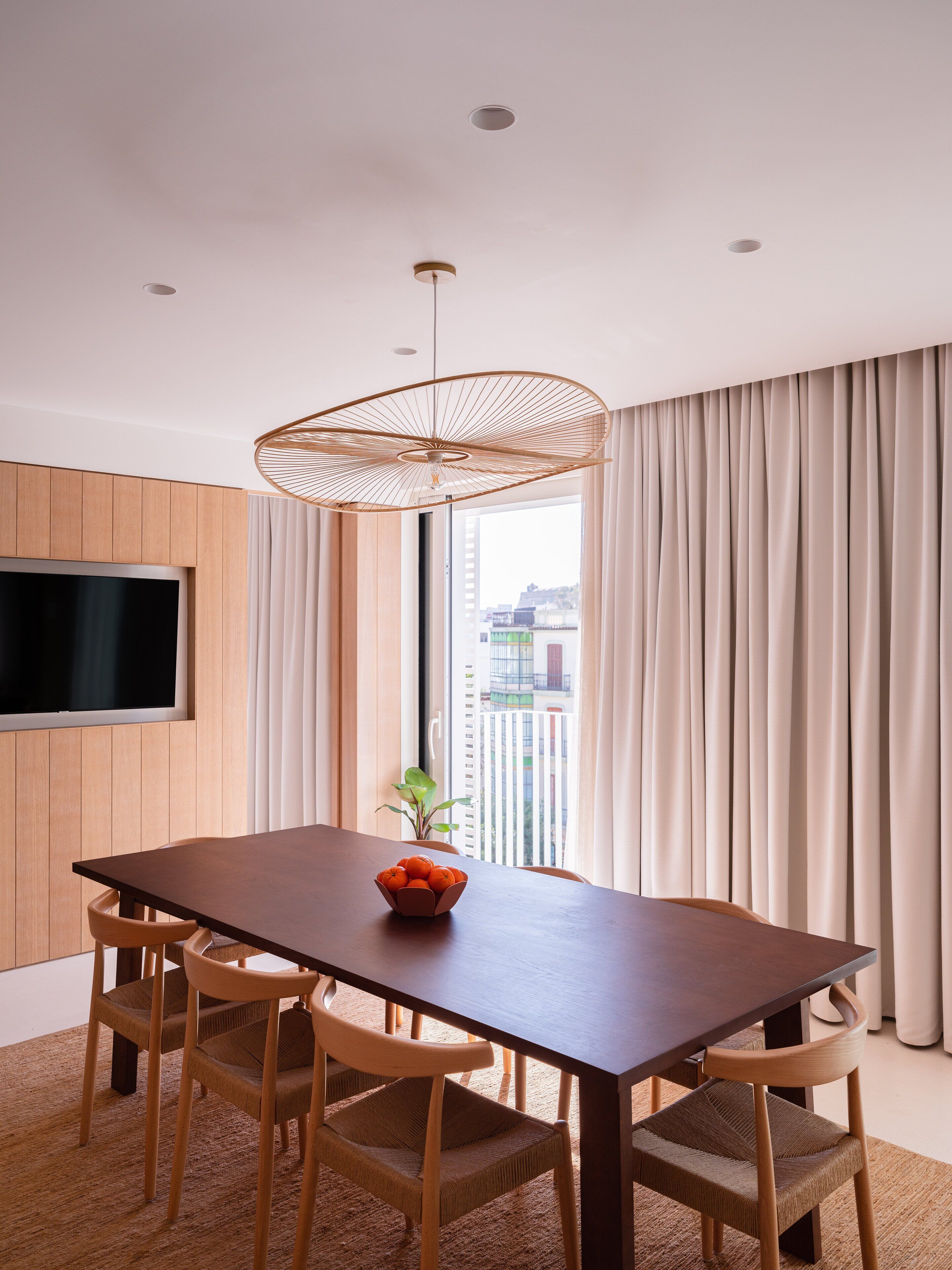 A modern dining area with a wooden table, four chairs, a woven pendant light, and a flat-screen TV mounted on the wall.