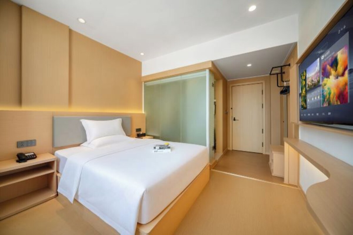 A modern hotel room with a large bed, bedside tables, a flat-screen TV, and a glass partition wall.