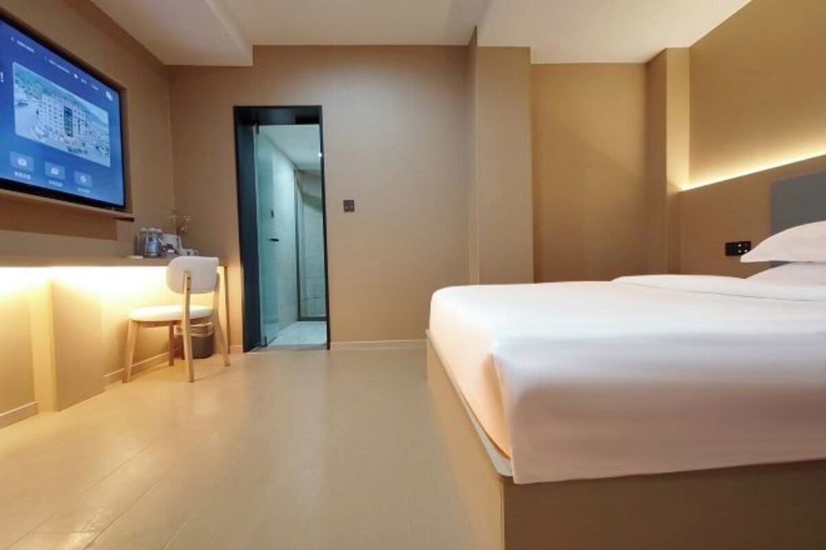 A hotel room with a bed, a desk, a chair, a TV, and a door leading to a hallway.