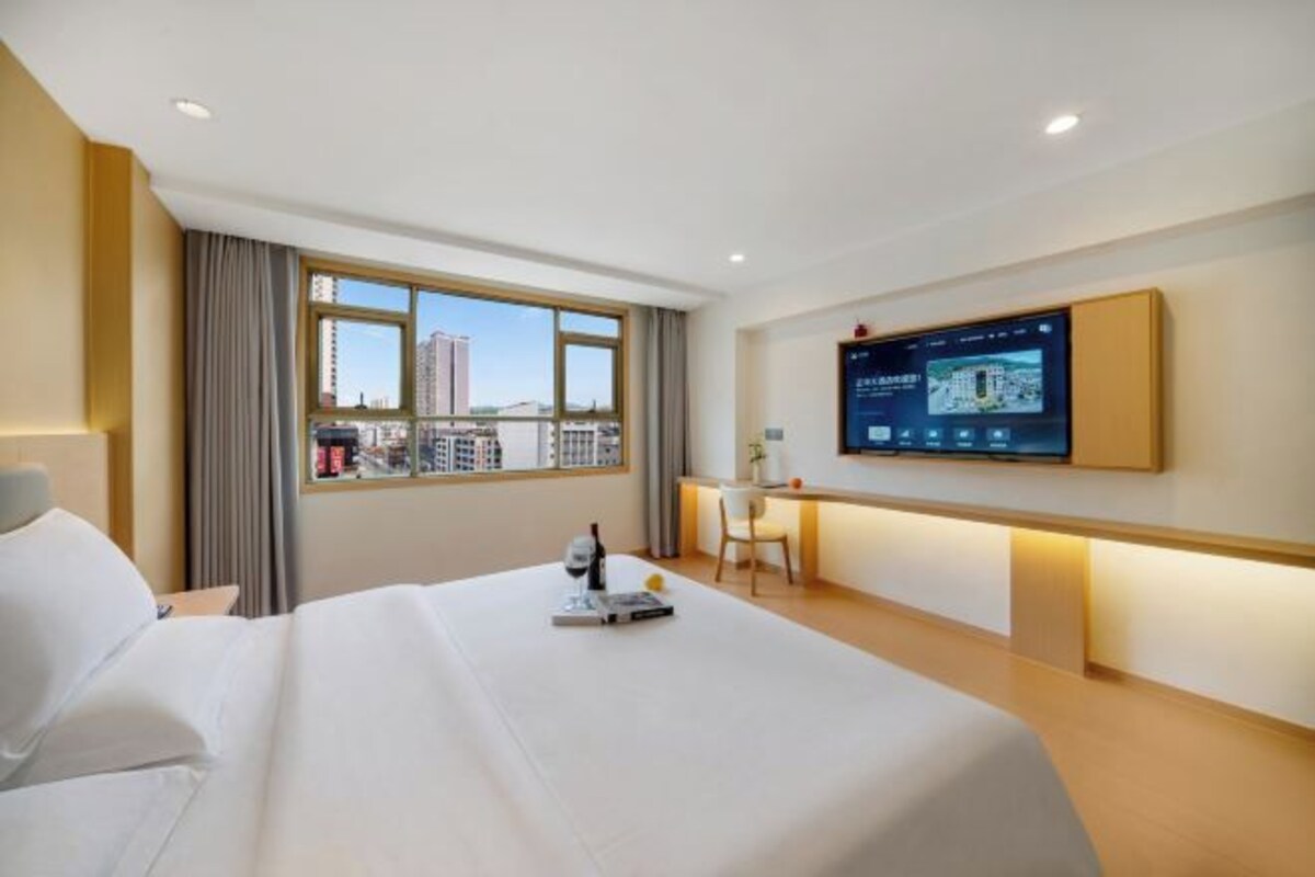 A hotel room with a large bed, a desk, a chair, a TV mounted on the wall, and a window with a city view.