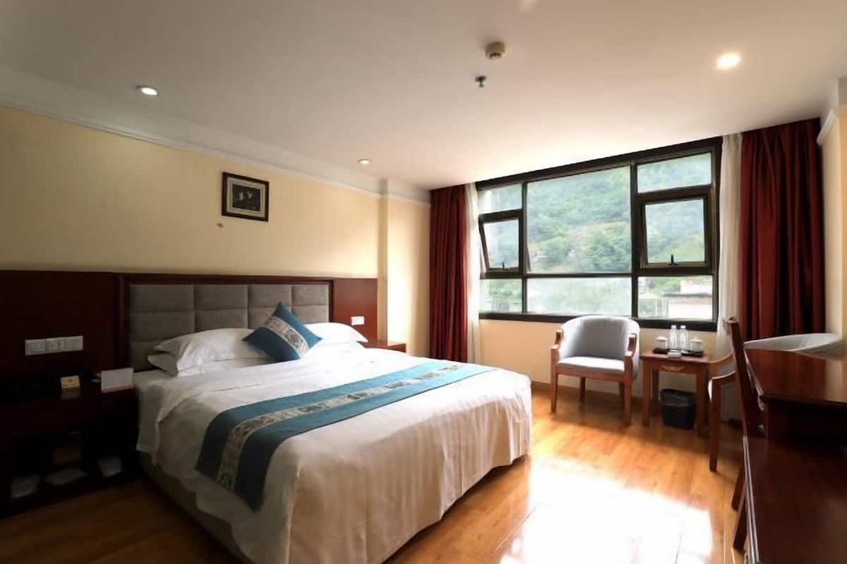 A hotel room with a large bed, a desk with a chair, a window with curtains, and a view of greenery outside.
