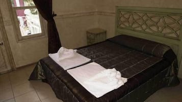 Una cama individual con un cabecero oscuro y una colcha blanca en una habitación con suelo de baldosas y una mesita de noche pequeña.