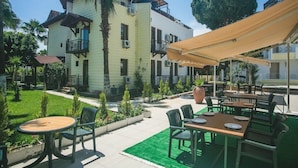 Un patio al aire libre con mesas y sillas, rodeado de un jardín cuidado y un edificio de dos pisos con balcones.