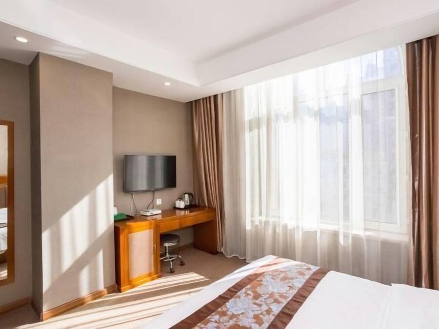 A hotel room with a bed, a desk, a television, and a large window with curtains.