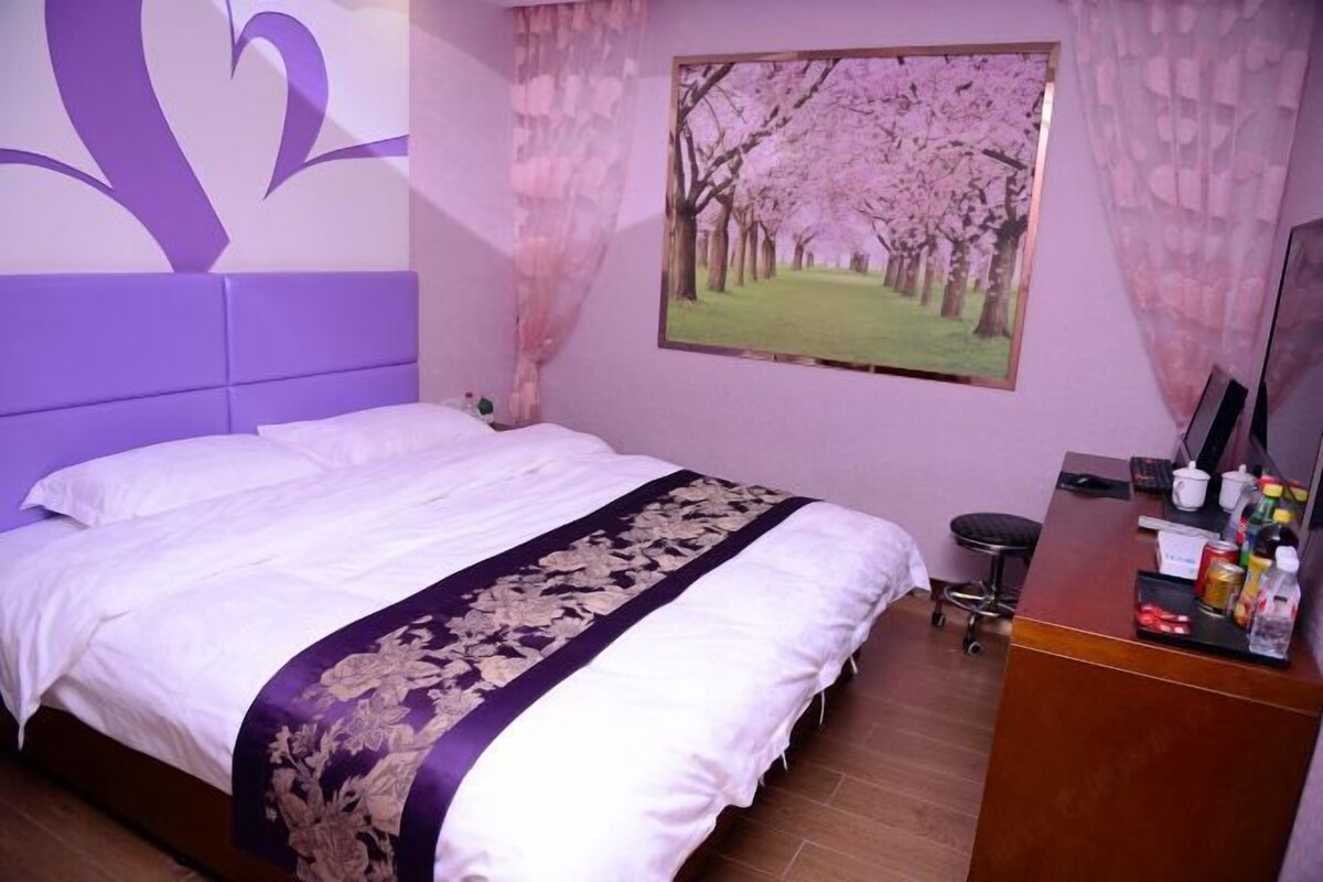 A bedroom with a bed, a desk with a chair, a painting of cherry blossoms, and a purple wall design.
