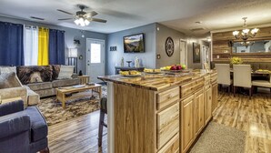 Una cocina moderna con una isla de madera, un comedor con una lámpara de araña y una sala de estar con sofá y cortinas.