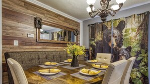 Un comedor con una mesa de madera dispuesta para cuatro personas, rodeada de sillas acolchadas. Un espejo cuelga sobre la mesa, y un tapiz con temática de osos adorna la pared.