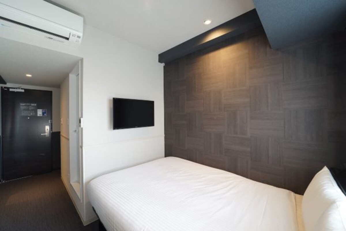 A hotel room with a bed, a flat-screen TV mounted on the wall, and a wooden-patterned headboard.
