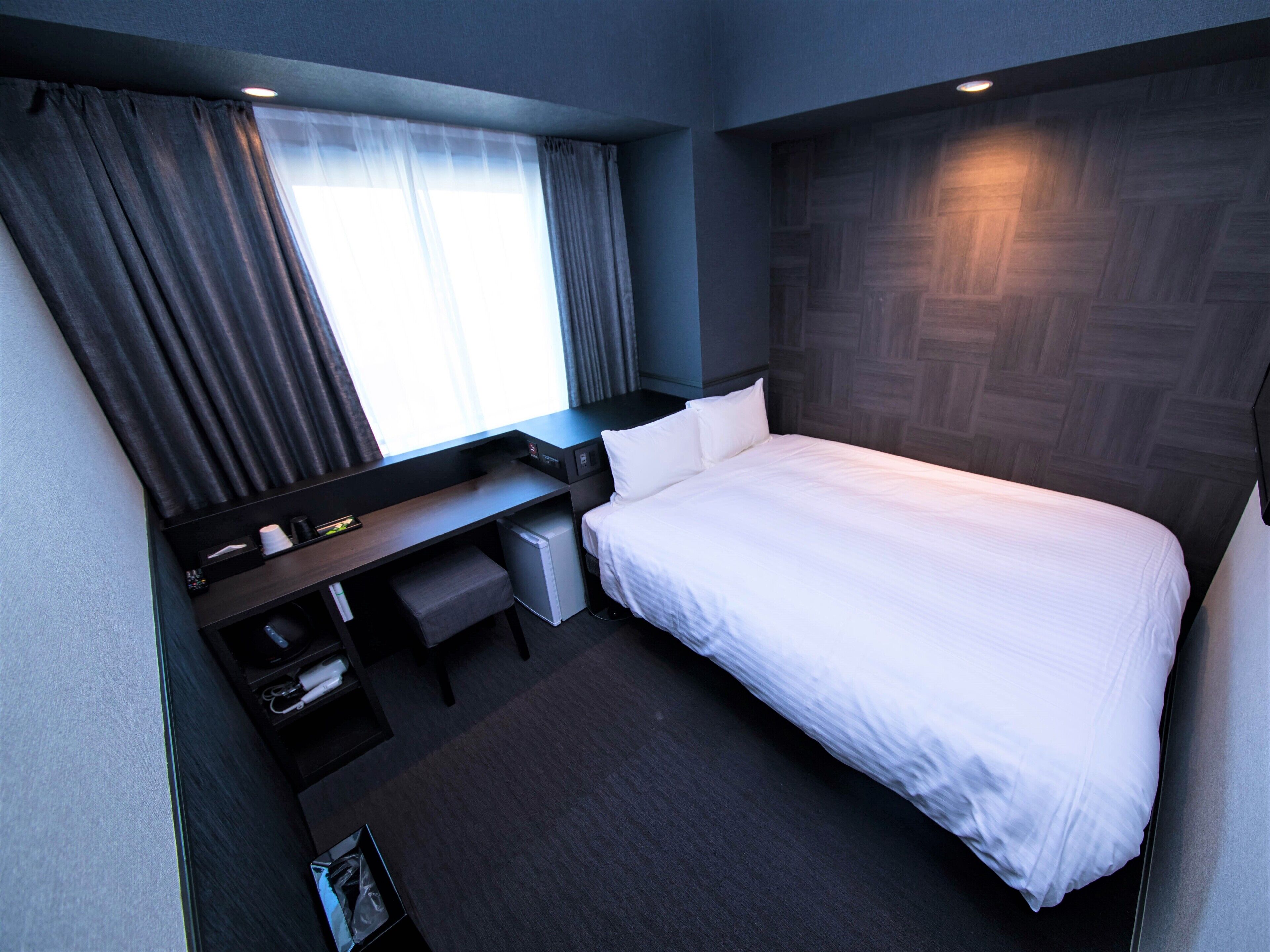 A modern hotel room with a neatly made bed, a desk with a chair, a small refrigerator, and a window with curtains.